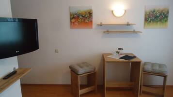 A small, tidy room with a flat-screen TV, two wooden chairs, a small table with a notebook, and two wall-mounted shelves with paintings.