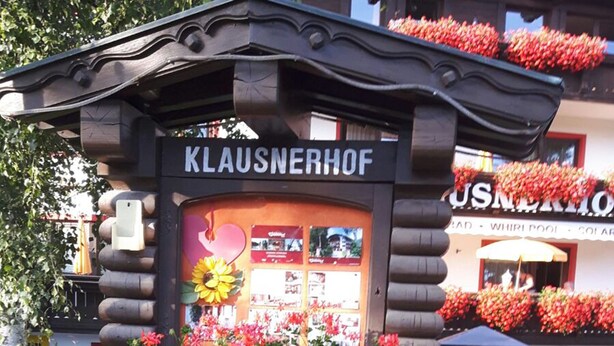 Un bâtiment de style chalet en rondins, appelé « KLAUSNERHOF », avec une enseigne, entouré de fleurs rouges éclatantes et de verdure.