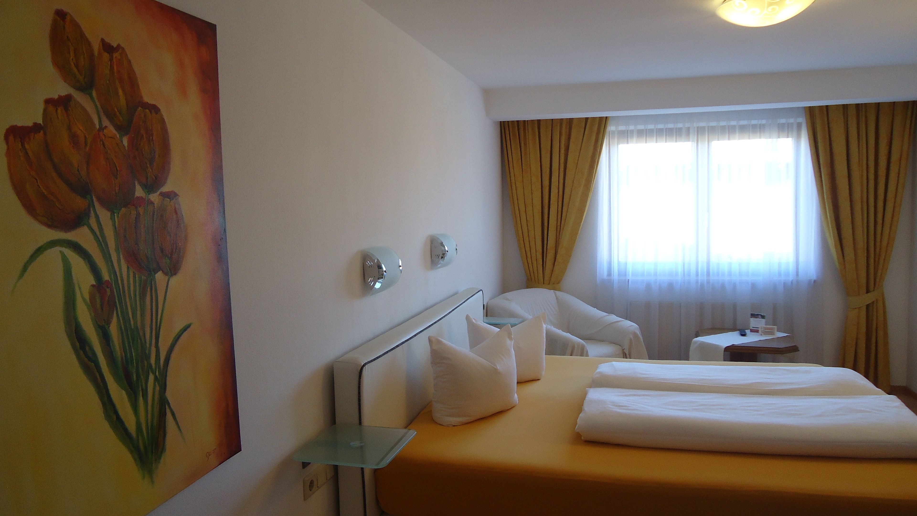 A hotel room with a bed, a chair, a small table, and a painting of tulips on the wall.