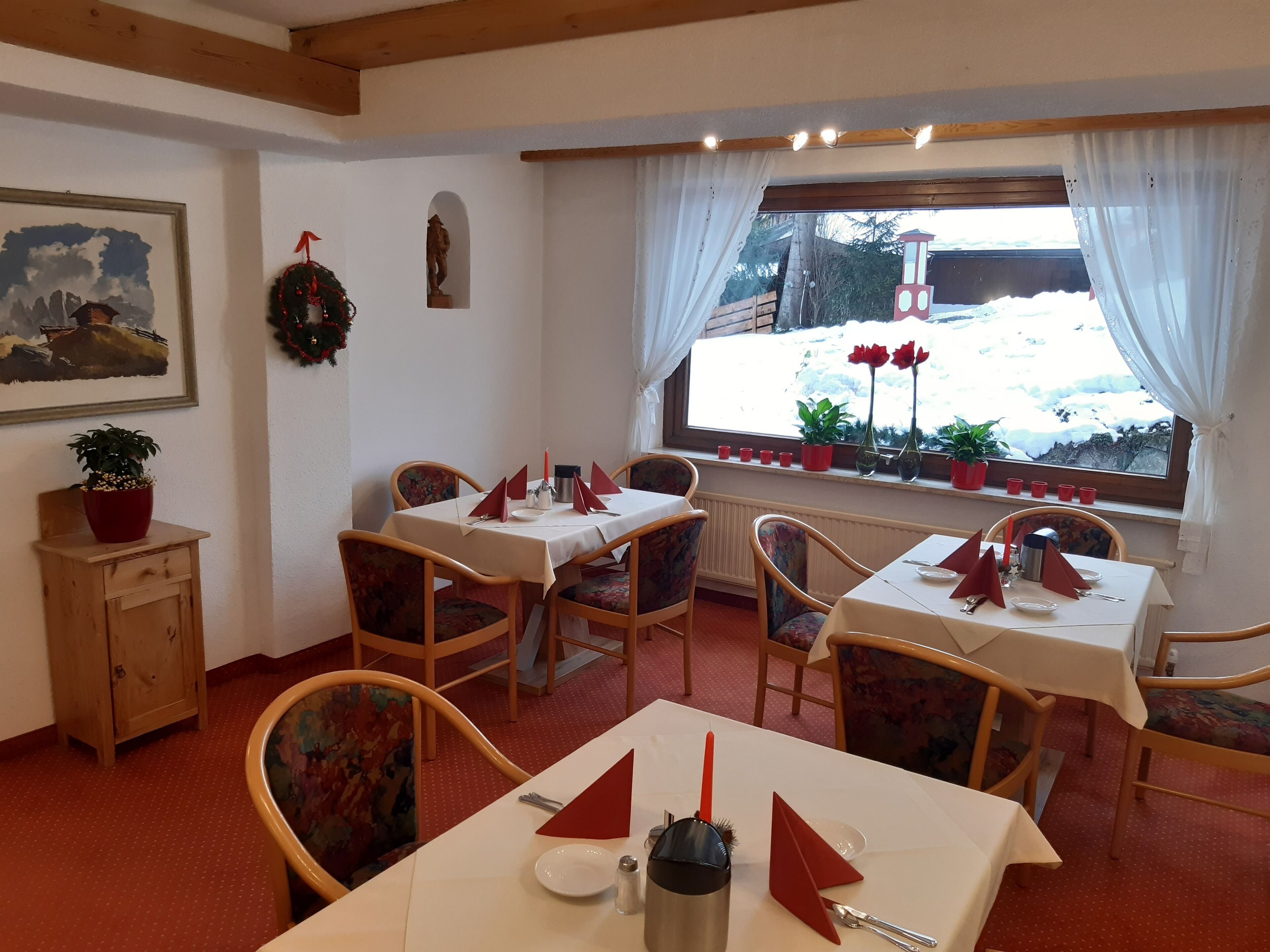 Un coin repas avec des tables préparées pour deux personnes, des chaises en bois, une fenêtre donnant sur un paysage enneigé et un petit meuble orné d’une plante.