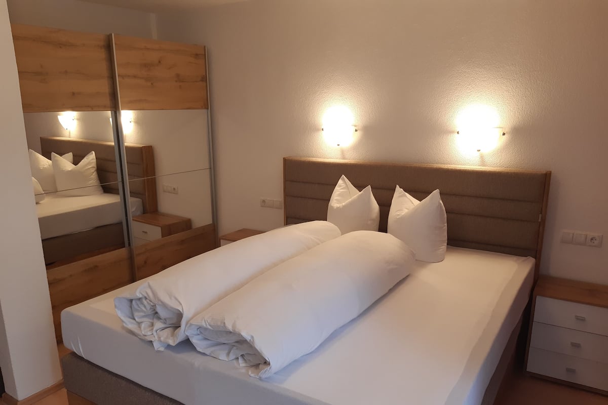 Un lit bien fait, avec du linge de lit blanc et des oreillers, une tête de lit en bois et des lampes fixées au mur.