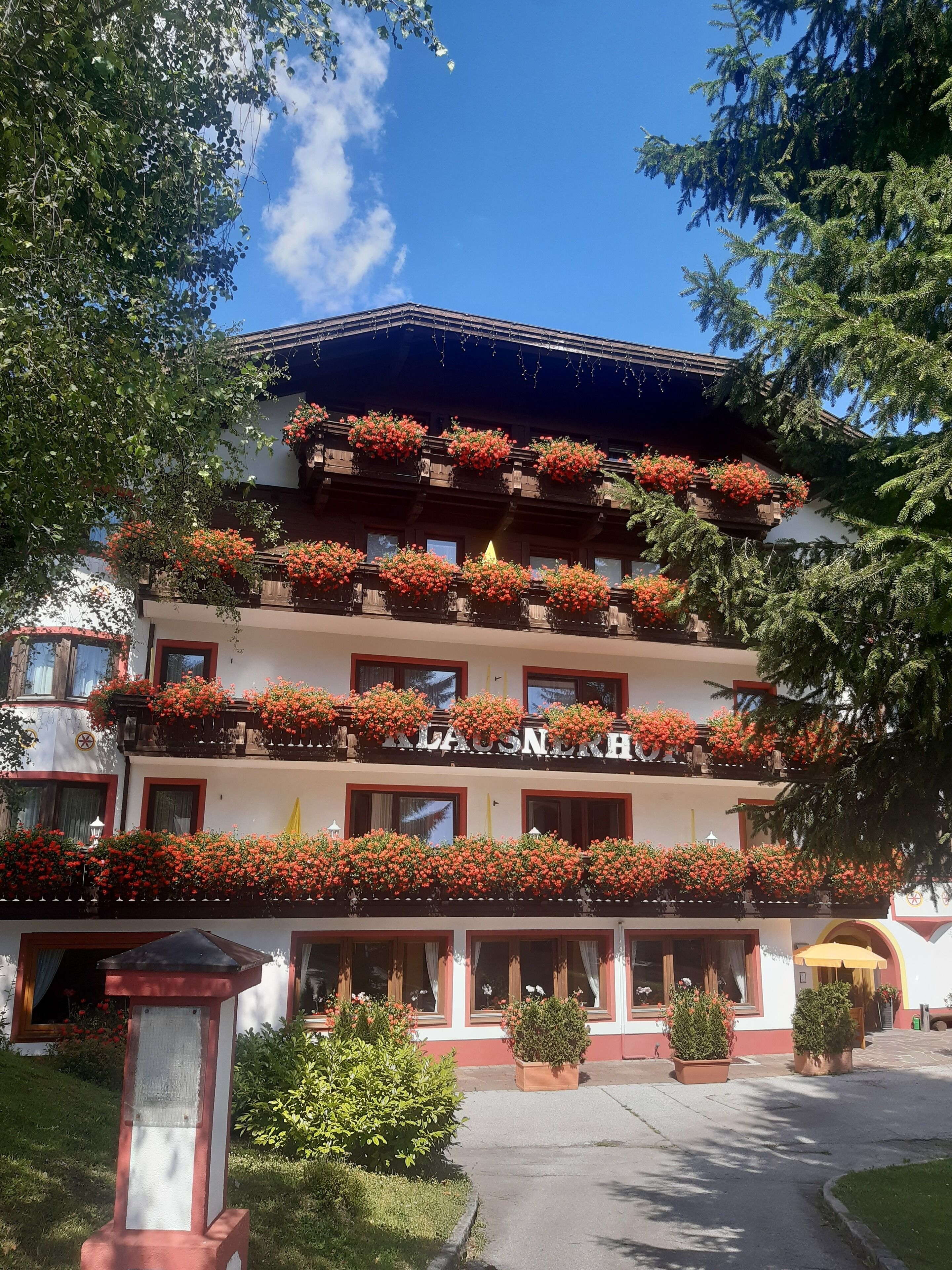 A multi-story hotel with balconies adorned with red flowers, a clear blue sky, and a well-maintained garden area.