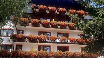 A multi-story hotel with balconies adorned with red flowers, a clear blue sky, and a well-maintained garden area.