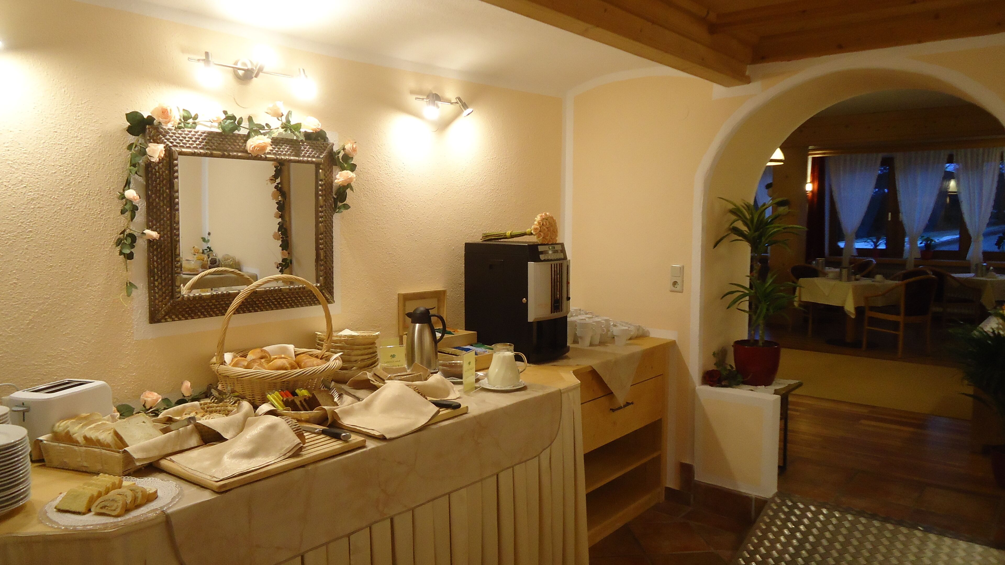 A breakfast bar with bread, pastries, and coffee equipment. There is a mirror with a floral wreath, a coffee machine, and a potted plant.