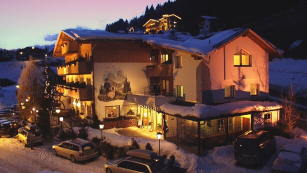 A multi-story hotel with a snow-covered roof, a ski slope in the background, and several parked cars in the foreground.