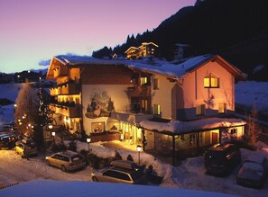 A multi-story hotel with a snow-covered roof, a ski slope in the background, and several parked cars in the foreground.