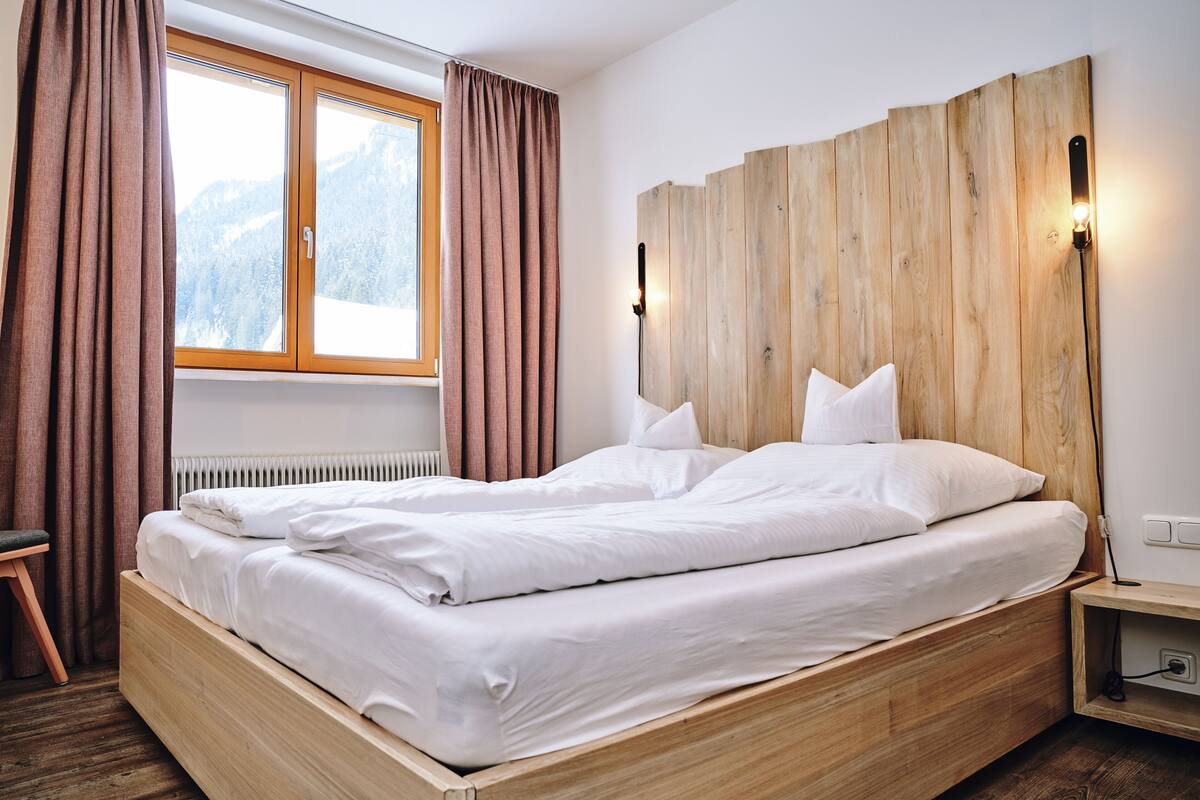 Una cama de madera con ropa de cama blanca, una ventana con cortinas y una mesita de noche con una lámpara.