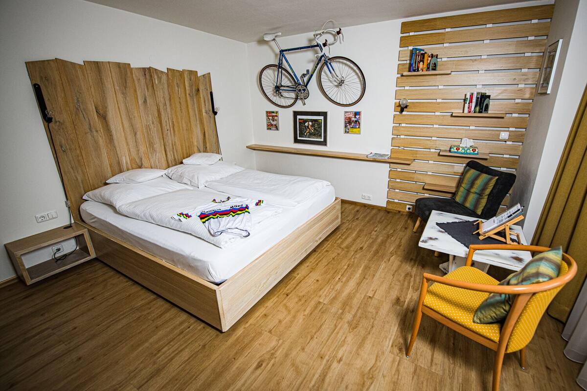 Un dormitorio con una cama de madera, una bicicleta colgada en la pared, una pequeña mesa con una silla y un estante de madera con libros y adornos.