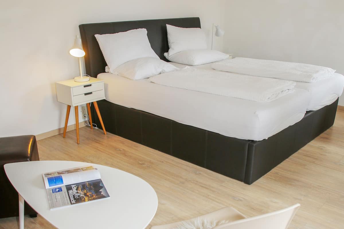 A neatly arranged bedroom with a bed, bedside table, lamp, chair, and a small table with a magazine.