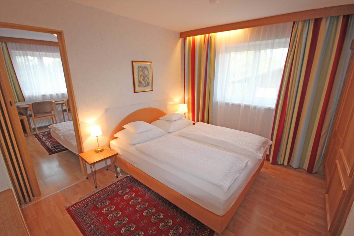 A hotel room with a large bed, a bedside table with a lamp, a chair, a small table, and a red patterned rug.