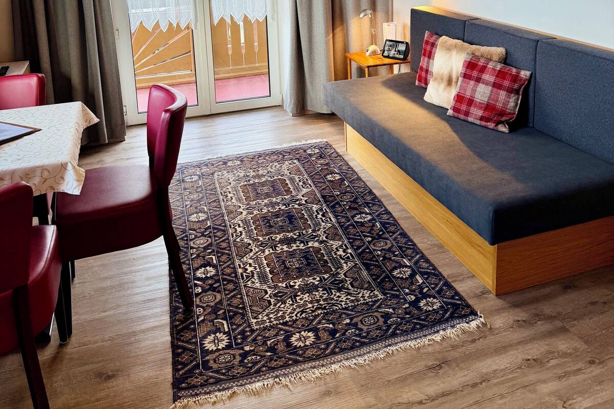 A cozy living area with a sofa, a dining table with chairs, a patterned rug, and a large window with curtains.