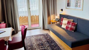 A cozy living area with a sofa, a dining table with chairs, a patterned rug, and a large window with curtains.