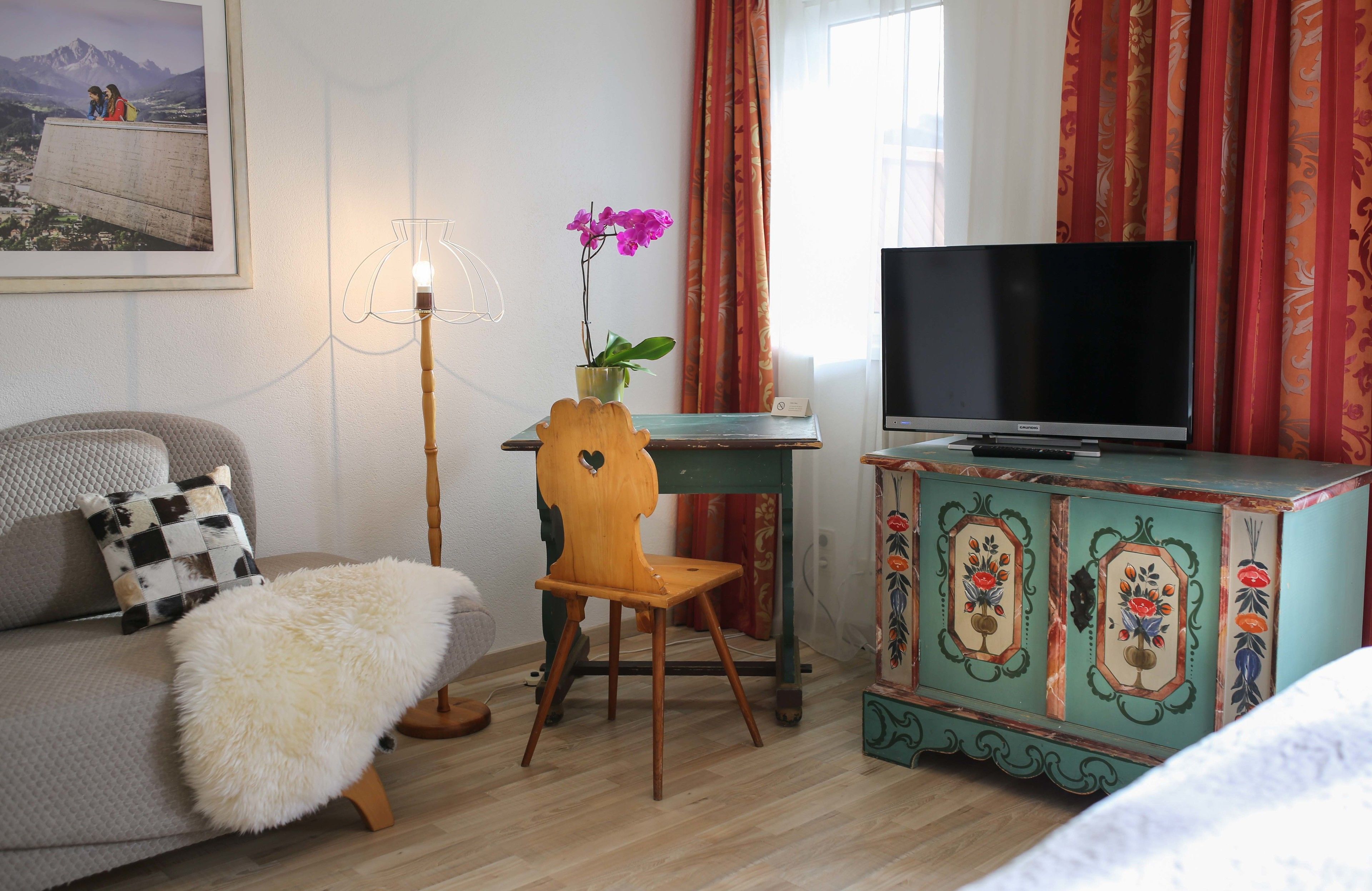 Habitación de hotel con sofá, una pequeña silla de madera, televisión, aparador decorativo y una orquídea en maceta.