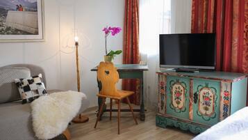 Habitación de hotel con sofá, una pequeña silla de madera, televisión, aparador decorativo y una orquídea en maceta.