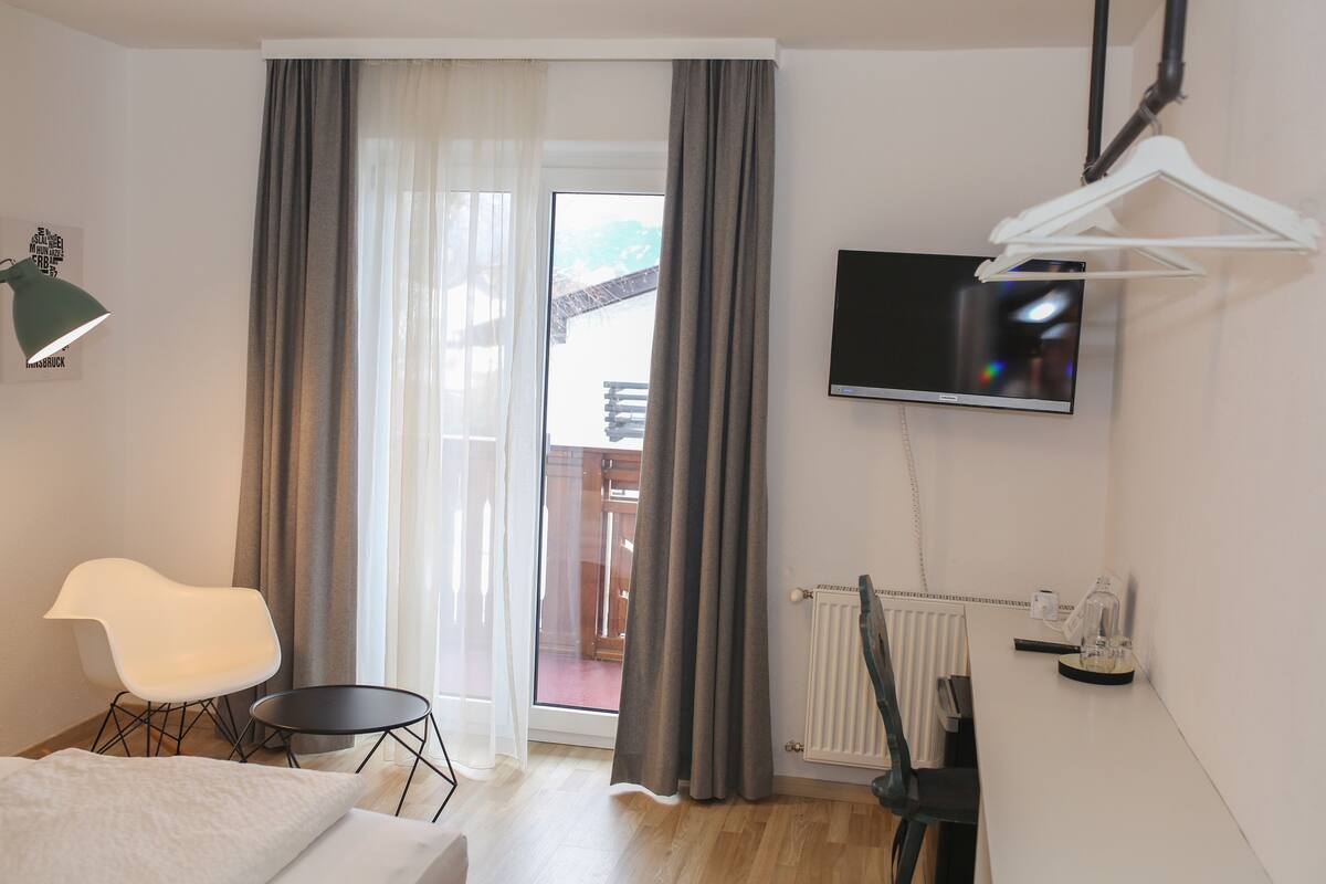 A modern hotel room with a bed, a white chair, a small table, a TV, and a view of the outside balcony.
