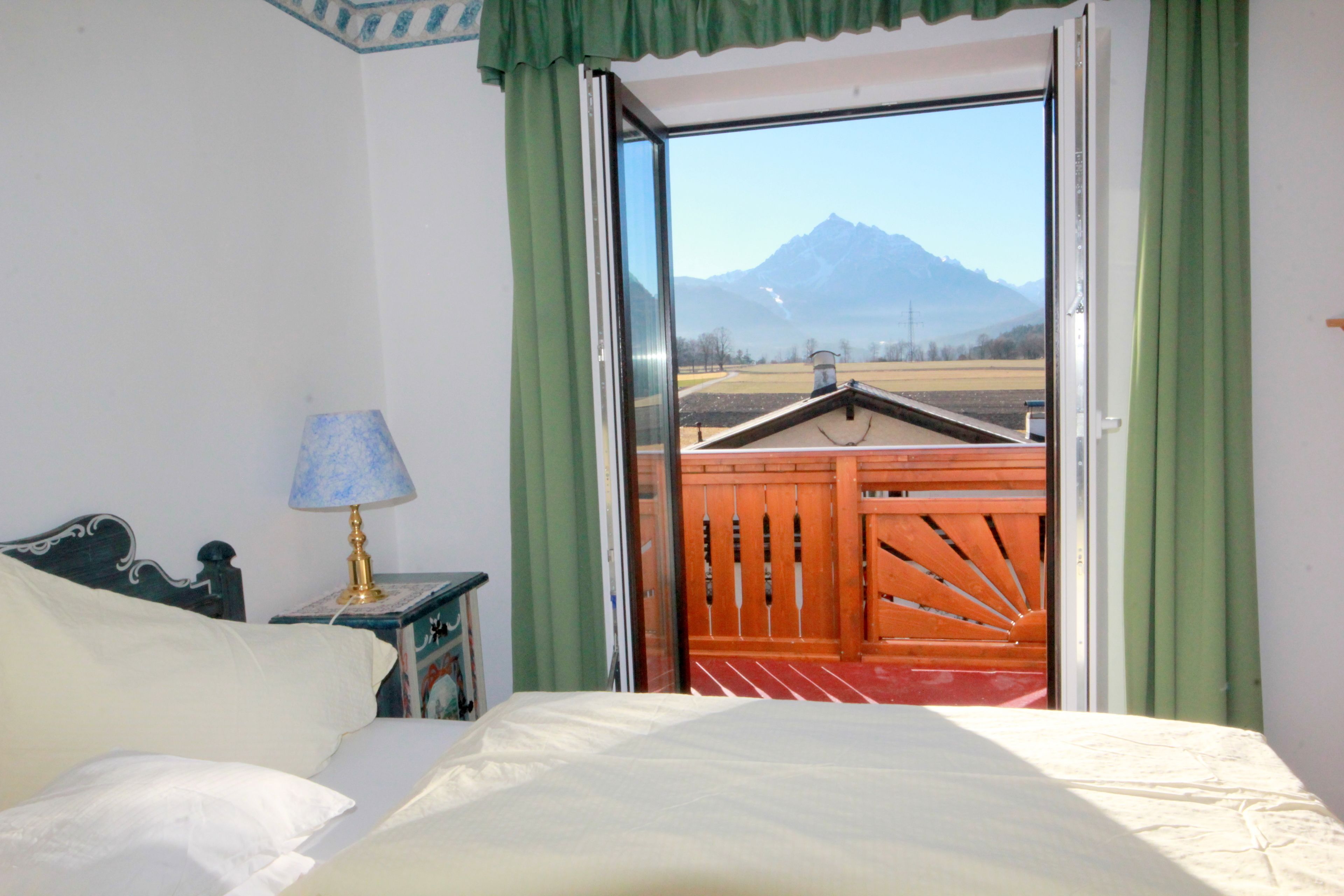 A hotel room with a bed, a nightstand with a lamp, and a view of mountains through an open door.