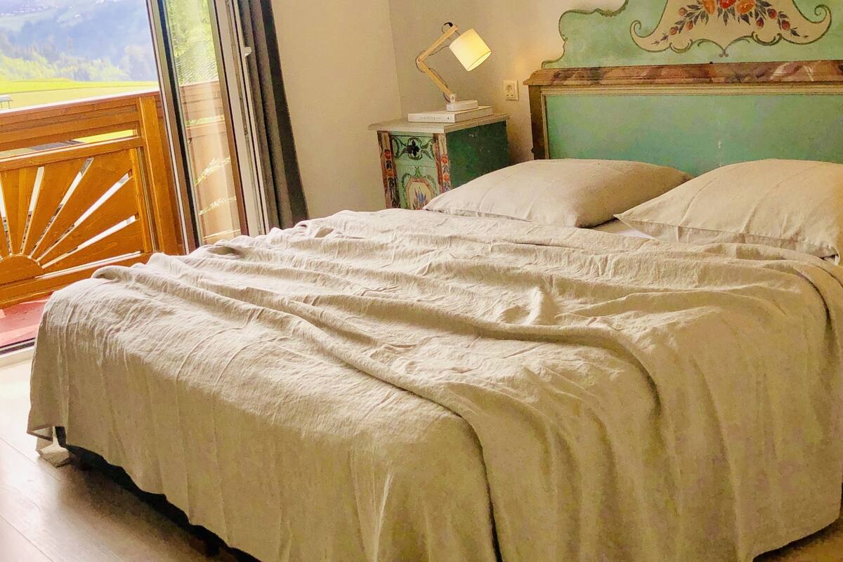 A bedroom with a large bed, a nightstand with a lamp, a decorative headboard, and a view of the outdoors through a sliding door.