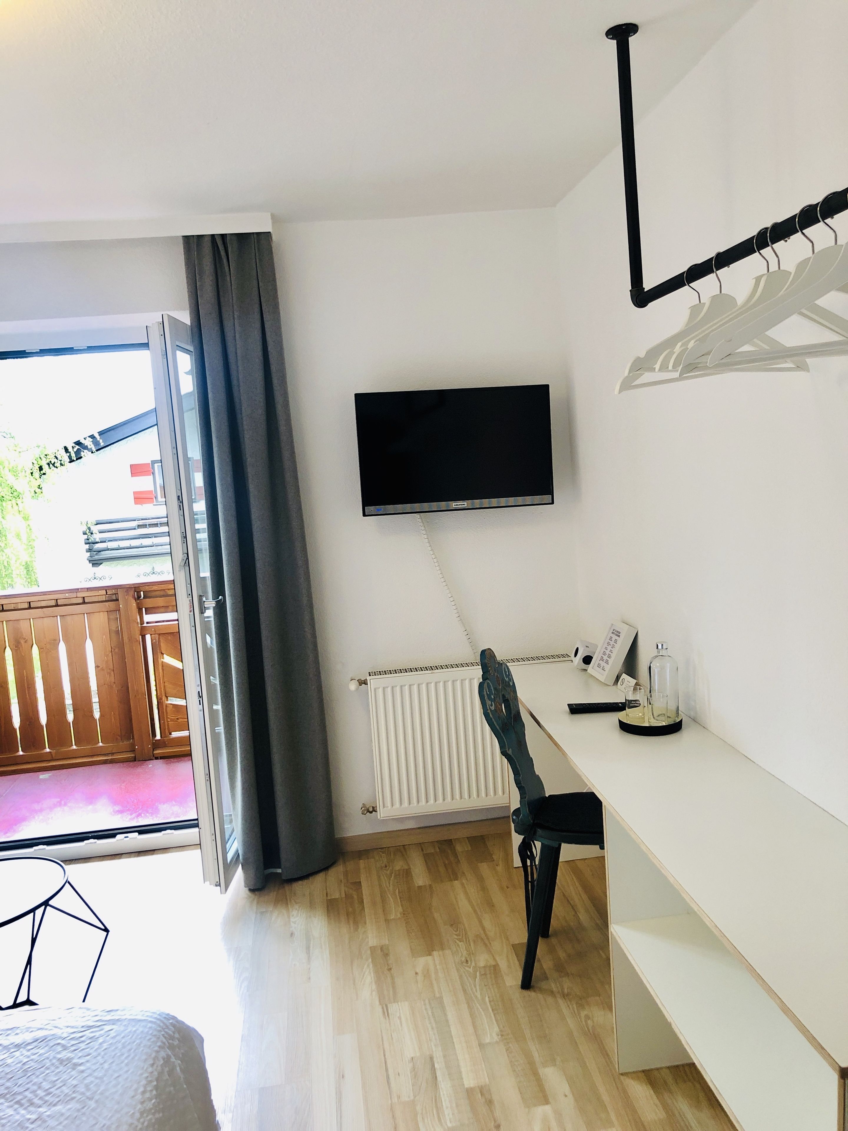 Una habitación con una cama, un escritorio con silla, un televisor montado en la pared, un radiador y una ventana con cortinas.