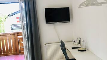 Una habitación con una cama, un escritorio con silla, un televisor montado en la pared, un radiador y una ventana con cortinas.