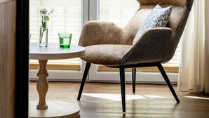 A room with a large window, a round wooden table with a vase and glass, and a brown upholstered in-arm chair with black legs. - Hotel Klingler (Eben am Achensee)