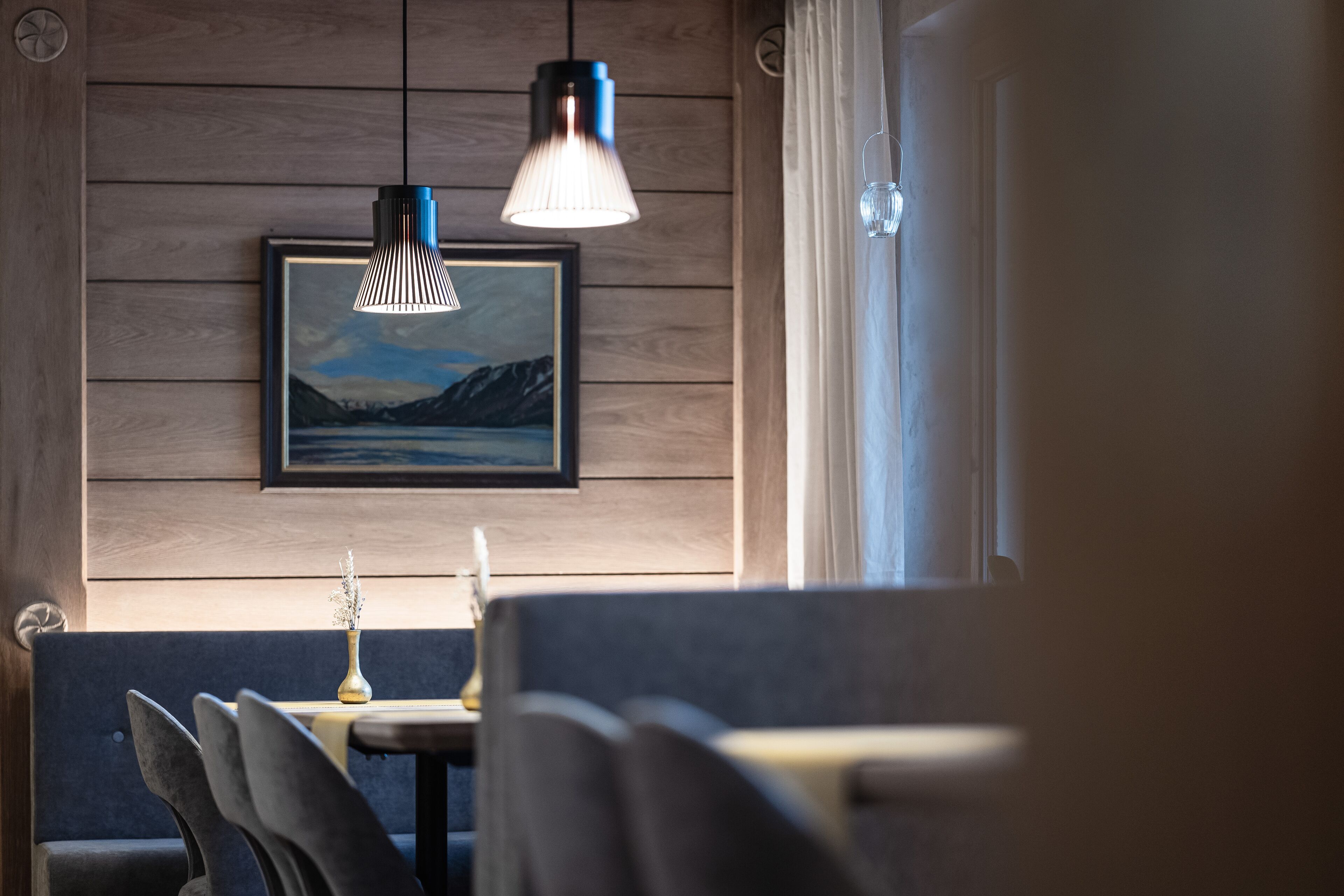Uma sala de jantar com parede de madeira, um quadro de paisagem emoldurado, uma mesa com um vaso e um lustre.