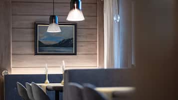 Uma sala de jantar com parede de madeira, um quadro de paisagem emoldurado, uma mesa com um vaso e um lustre.
