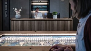 A reception desk with a person behind it, a vase with flowers, and a large illuminated map display in the foreground. - Hotel Klingler (Eben am Achensee)