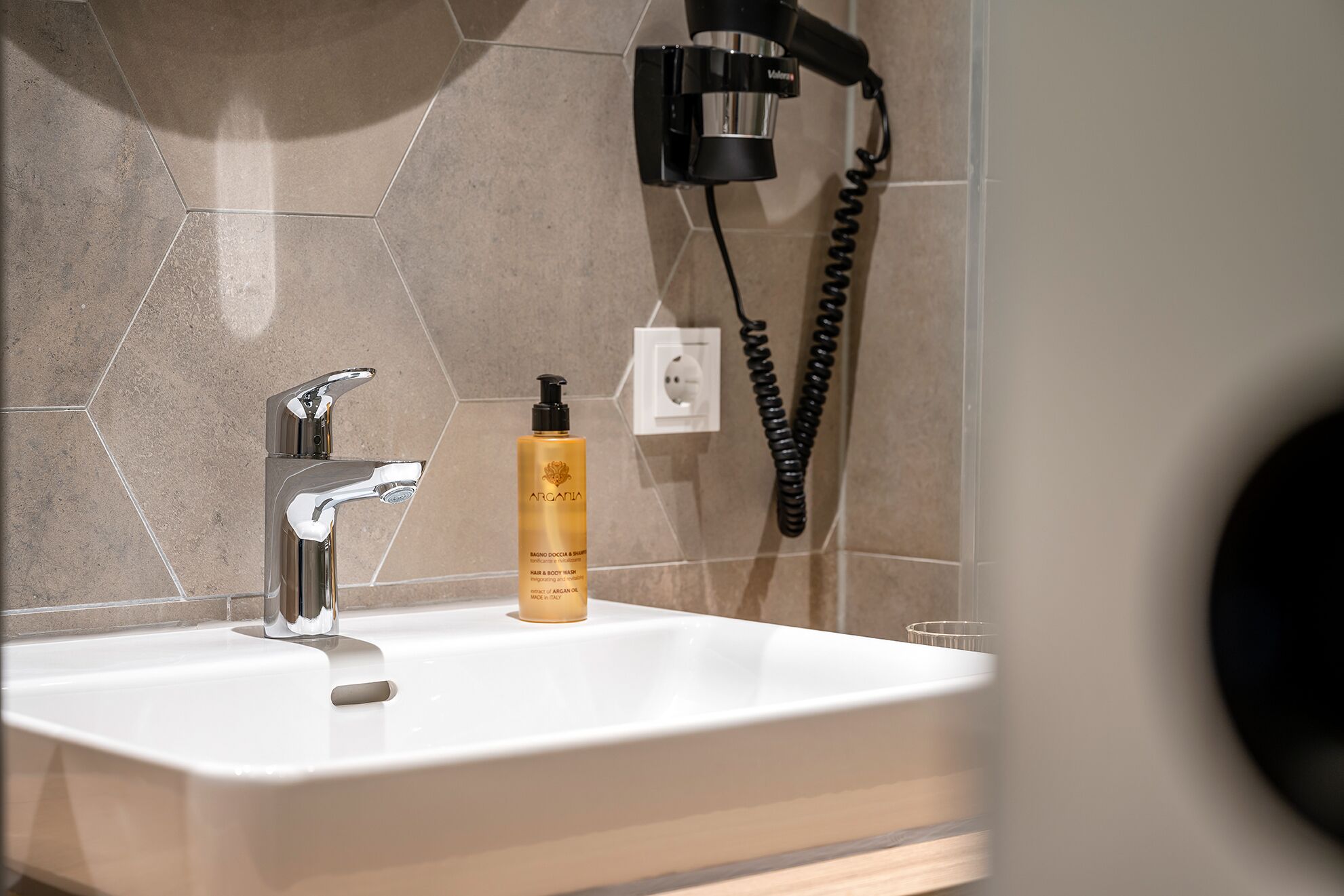 Ein Badezimmer mit einem weißen Waschbecken, einem modernen Wasserhahn, einer Flasche mit Ha- und Körperpflegeprodukten und einem schwarzen Fön, der an der Wand befestigt ist.