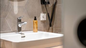 Ein Badezimmer mit einem weißen Waschbecken, einem modernen Wasserhahn, einer Flasche mit Ha- und Körperpflegeprodukten und einem schwarzen Fön, der an der Wand befestigt ist.