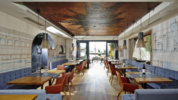 A restaurant with wooden tables and blue cushioned seating, a high ceiling with exposed beams, and a view of the street through large windows. - Hotel Klingler (Eben am Achensee)