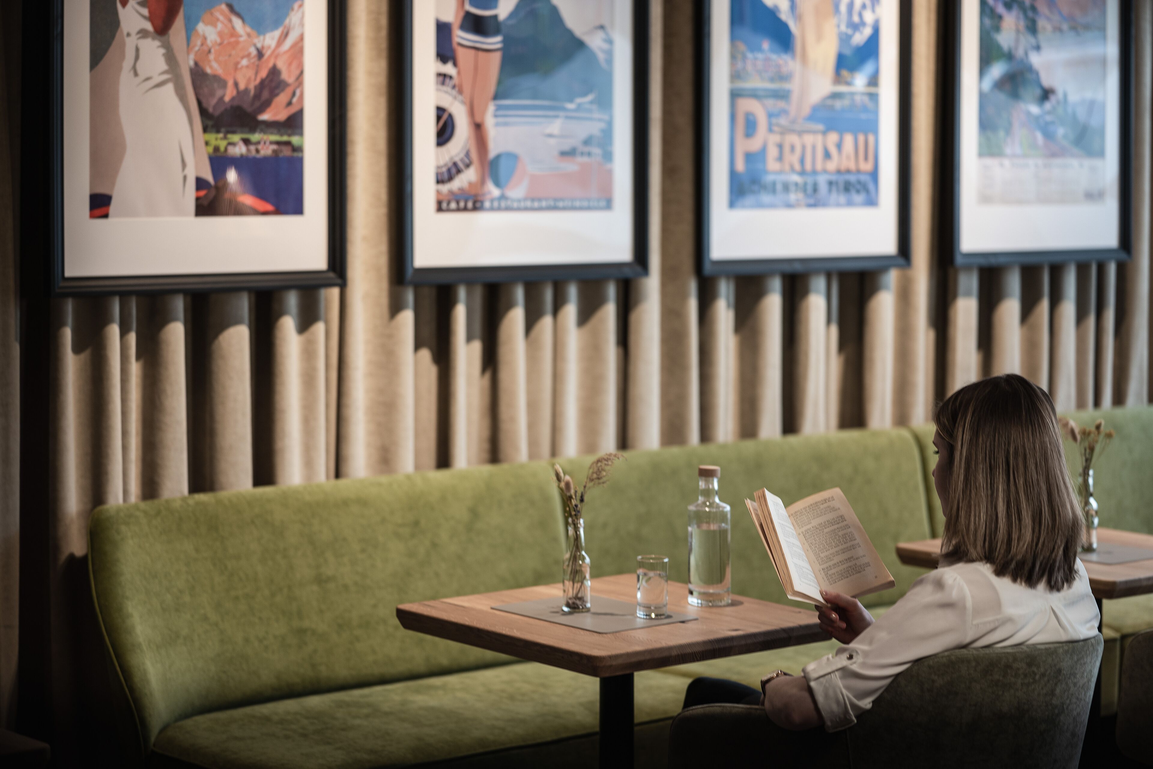 A person reading a book at a table with a green sofa and framed posters in the background.