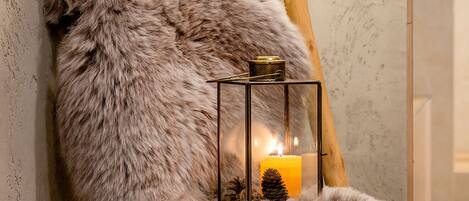 A cozy room with a wooden chair upholstered in faux fur, a table with magazines, and a floor lamp with a candle.