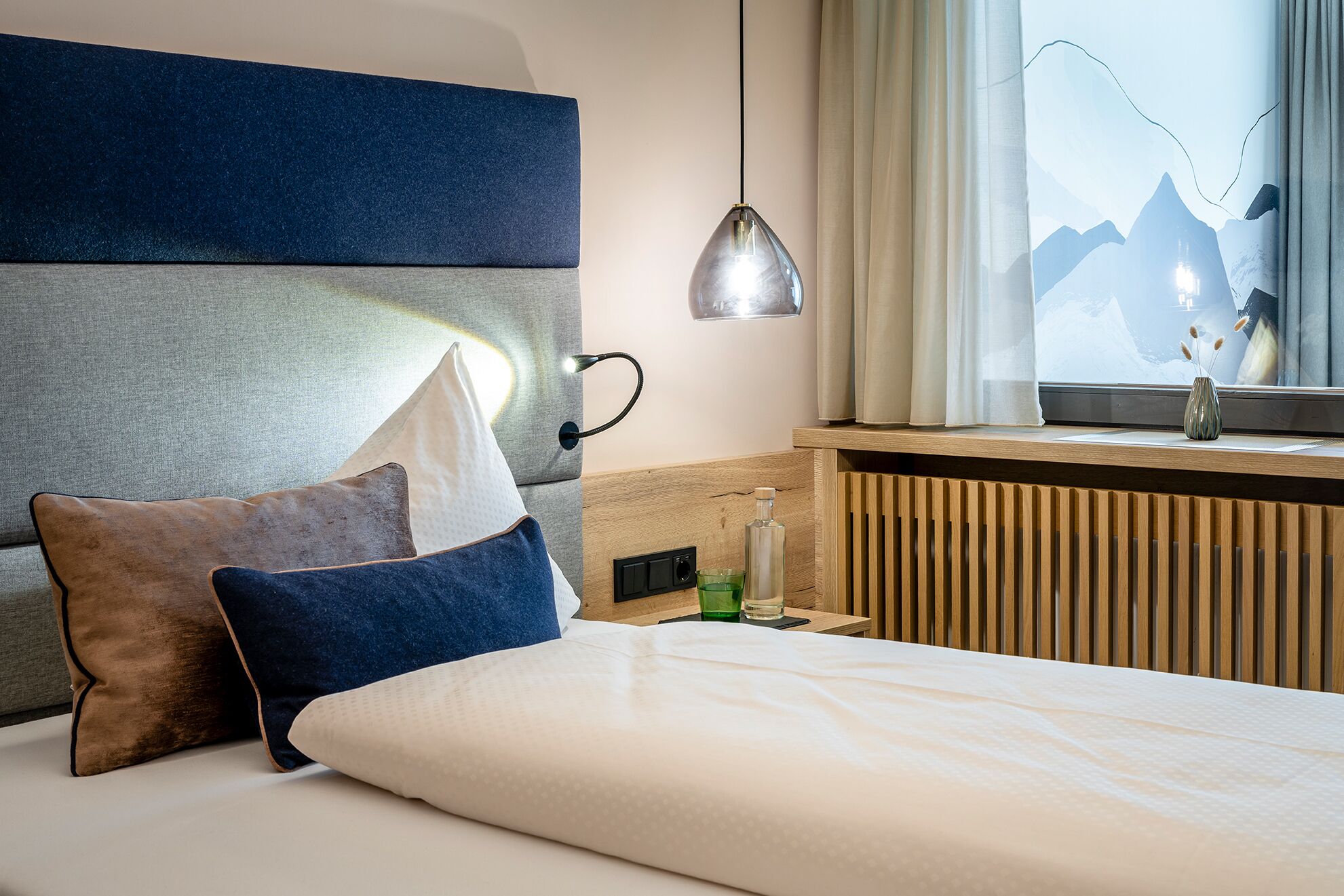 A hotel room with a bed, a nightstand, a lamp, a window with a view of mountains, and a radiator.