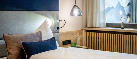 A hotel room with a bed, a nightstand, a lamp, a window with a view of mountains, and a radiator.