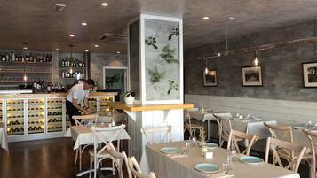A restaurant interior with wooden tables and chairs, a bar area with wine bottles, and a large decorative plant.