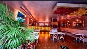 Ein Restaurant mit Holzboden, weißen Stühlen, einer Bar in der Mitte und einem großen Topfpflanzen.
