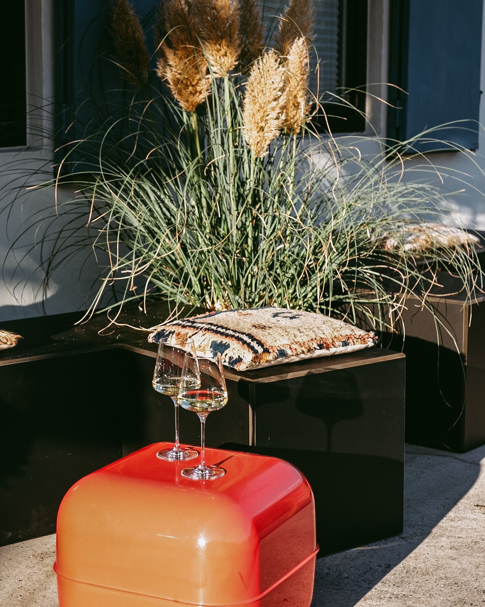 Un área de estar moderna al aire libre con un pouf rojo y una mesa de vidrio, rodeada de pastos altos y plumas de pampa.