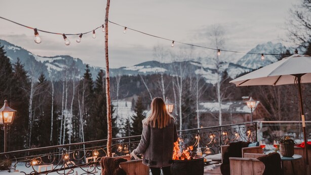 En person står på en balkon med udsigt over et sneklædt bjerglandskab med lyskæder og et bålsted.