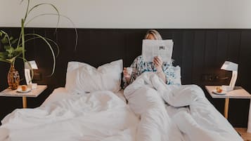 A person lying in bed reading a newspaper, with a plant on a bedside table and a lamp on the nightstand.