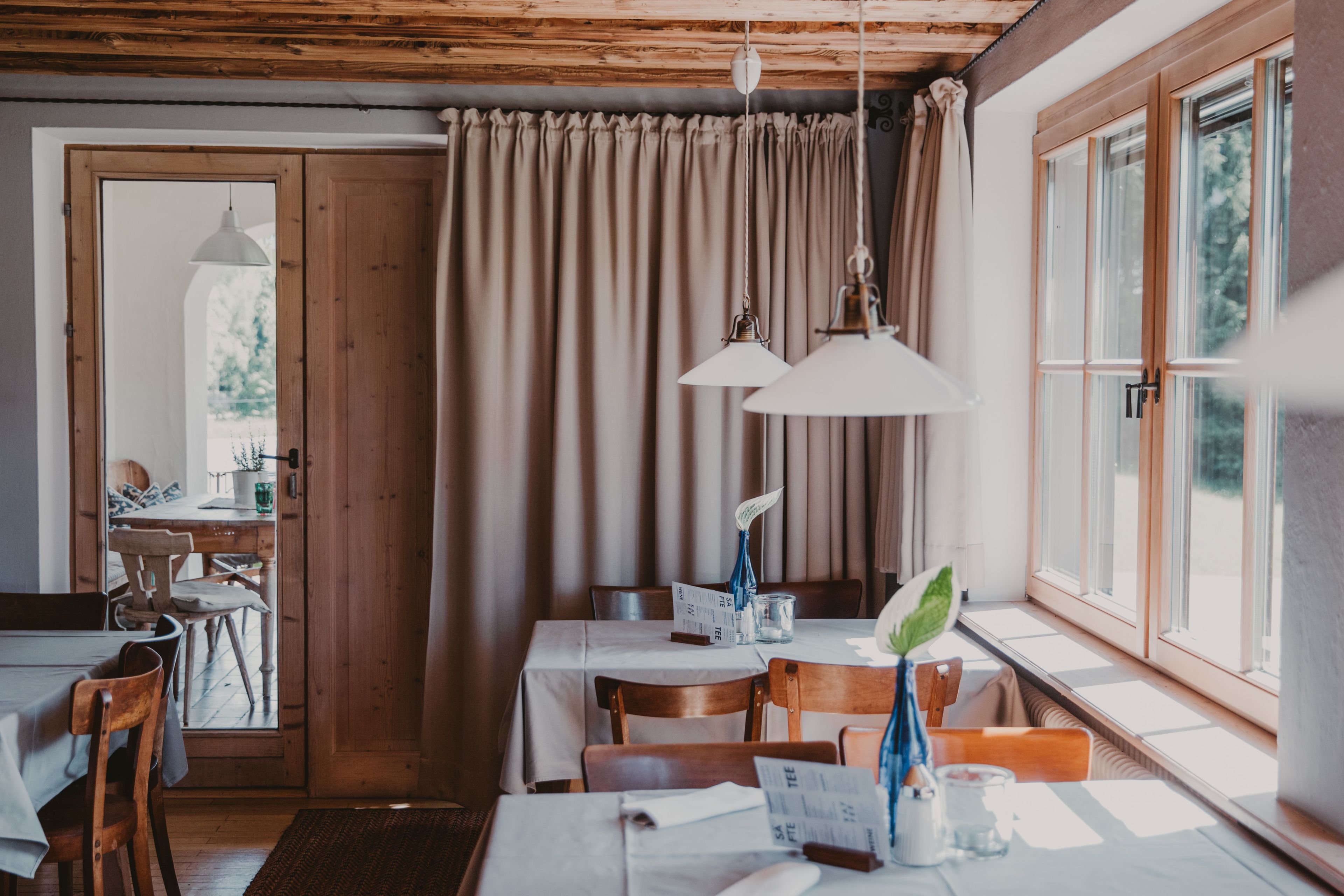 Un comedor con mesas y sillas de madera, una ventana con cortinas y lámparas colgantes.