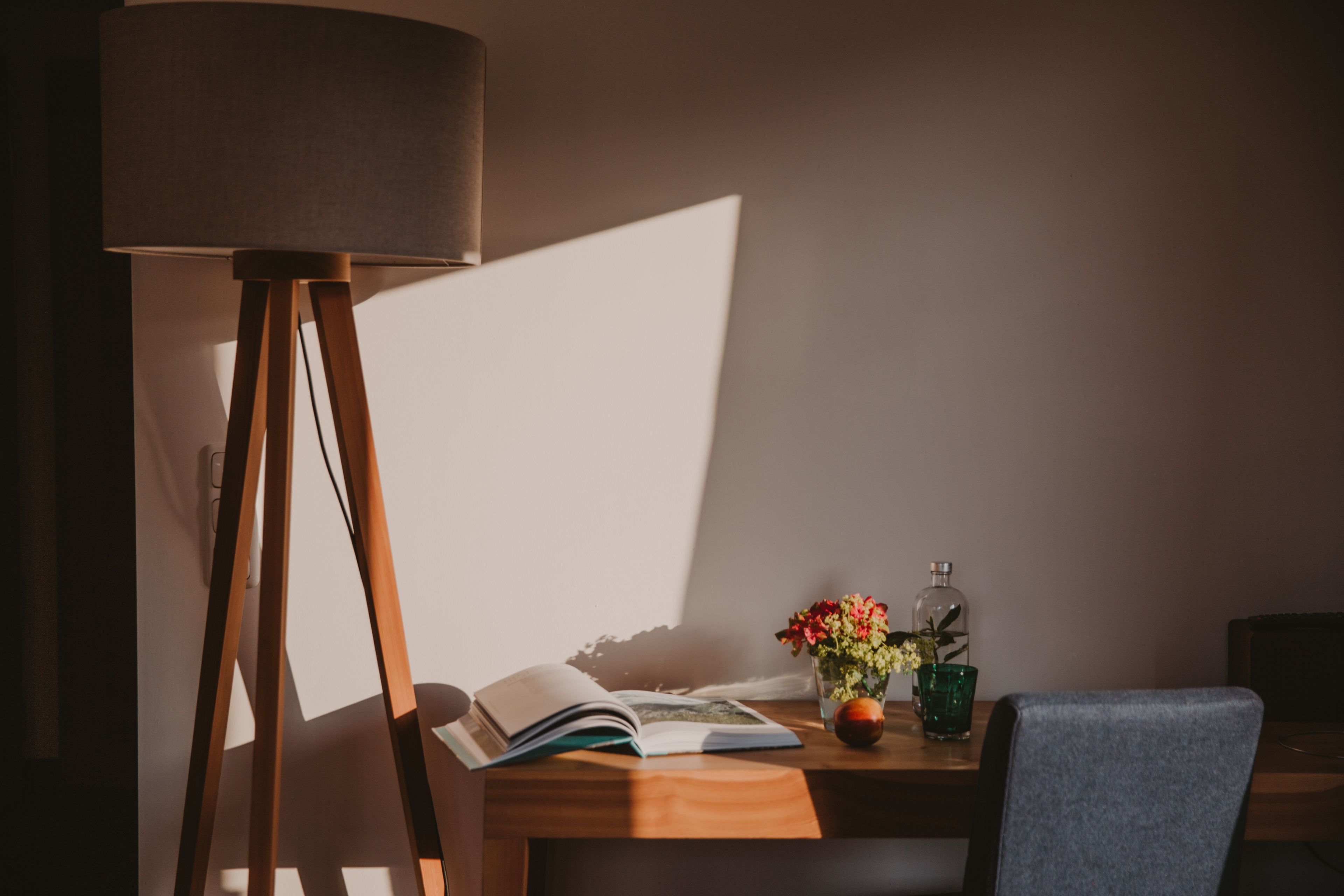 A wooden desk with a lamp, an open book, a vase with flowers, and a bottle.
