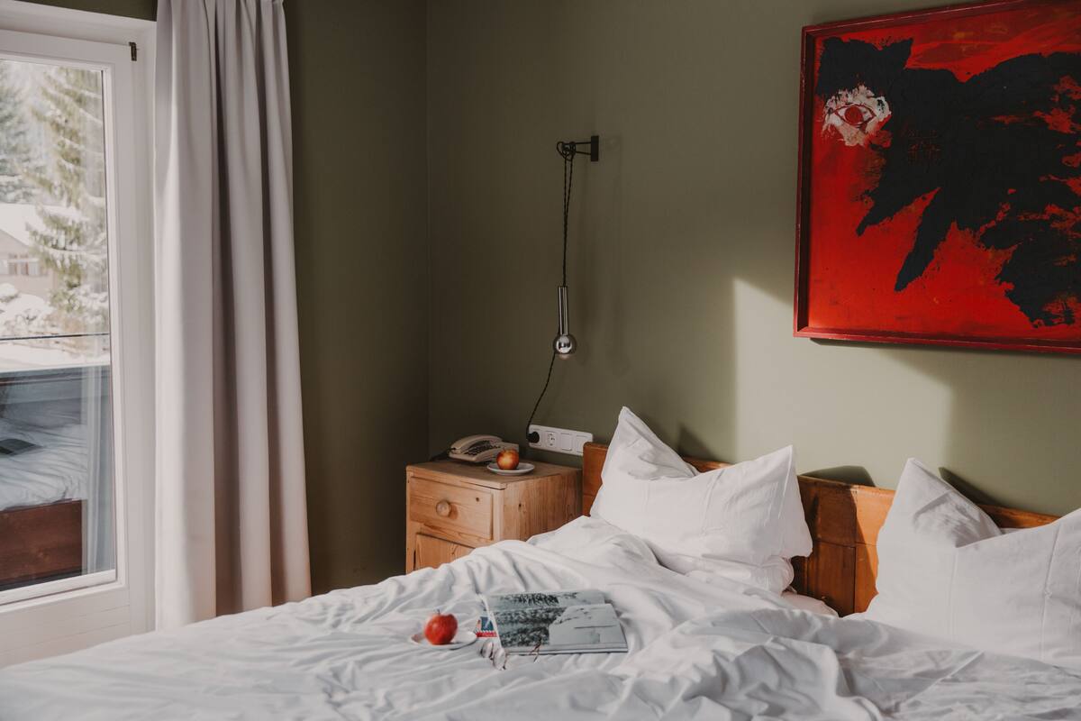 Habitación de hotel con una cama, una mesita de noche con un teléfono y una manzana, una ventana con cortinas y un cuadro abstracto rojo en la pared.