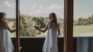 En person i en hvid kjole står ved et vindue med udsigt til et bjerglandskab.