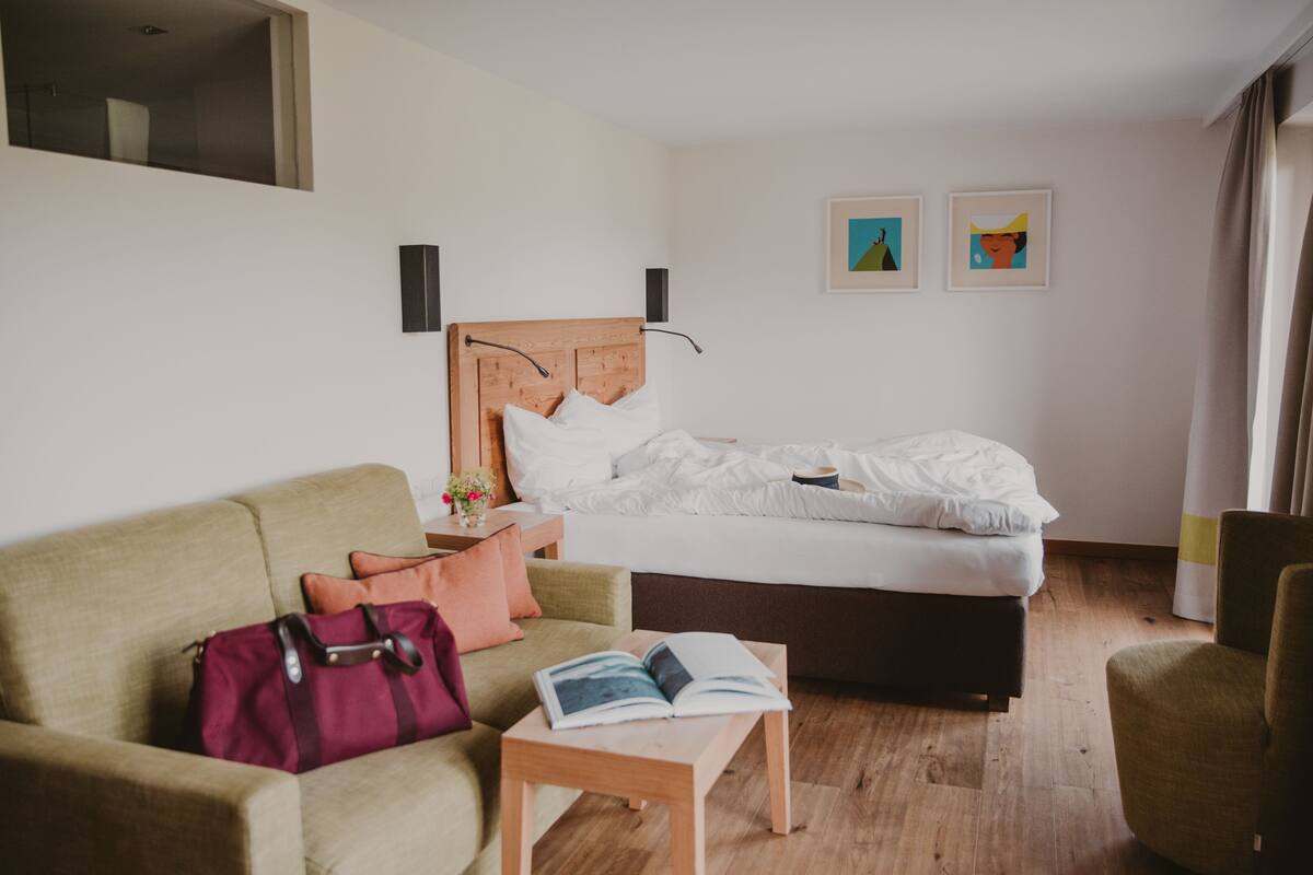 Habitación de hotel con cama, sofá, una mesa con un libro, una maleta y dos cuadros en la pared.