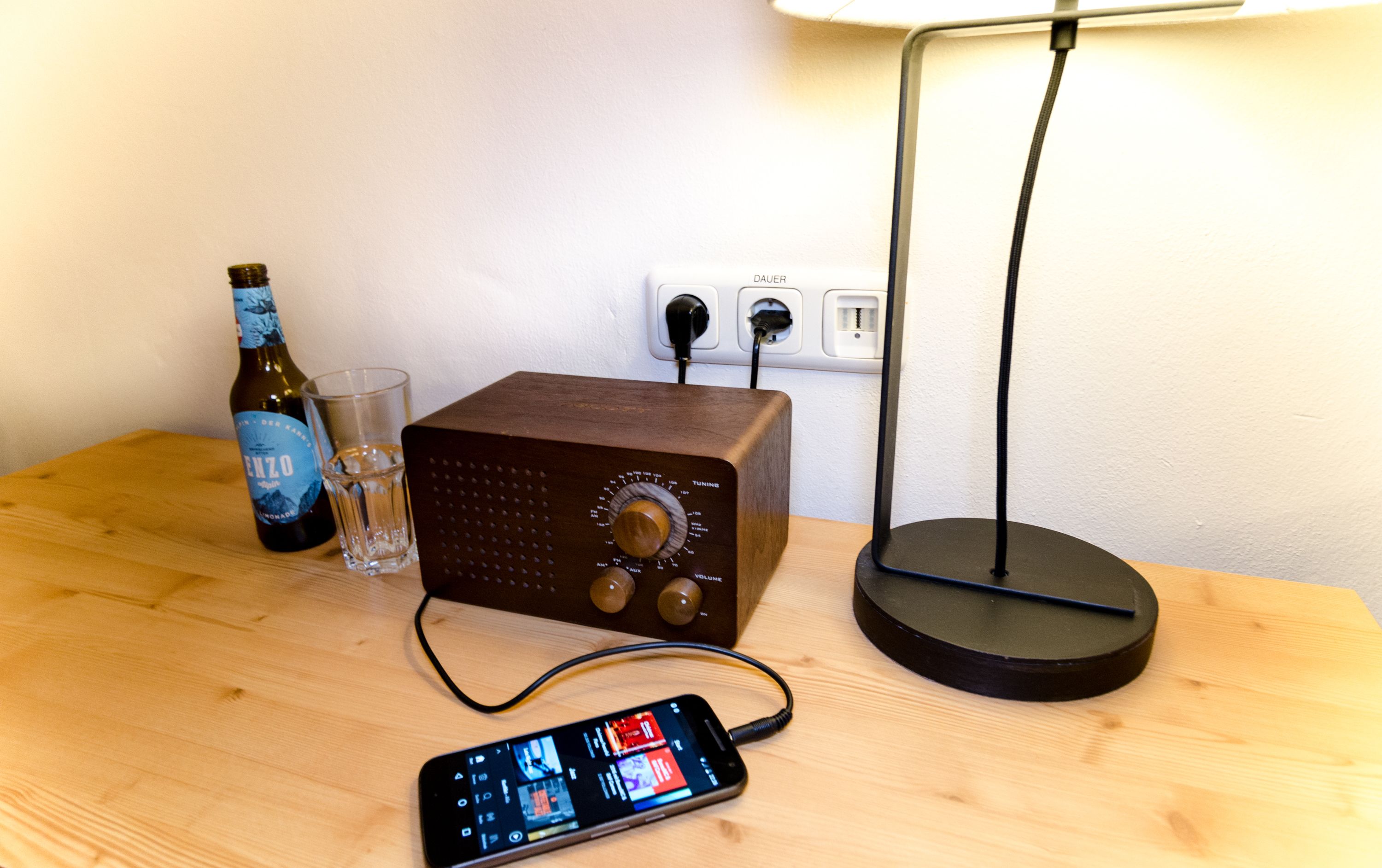 Uma mesa de madeira com uma garrafa, um copo, um rádio e um smartphone.