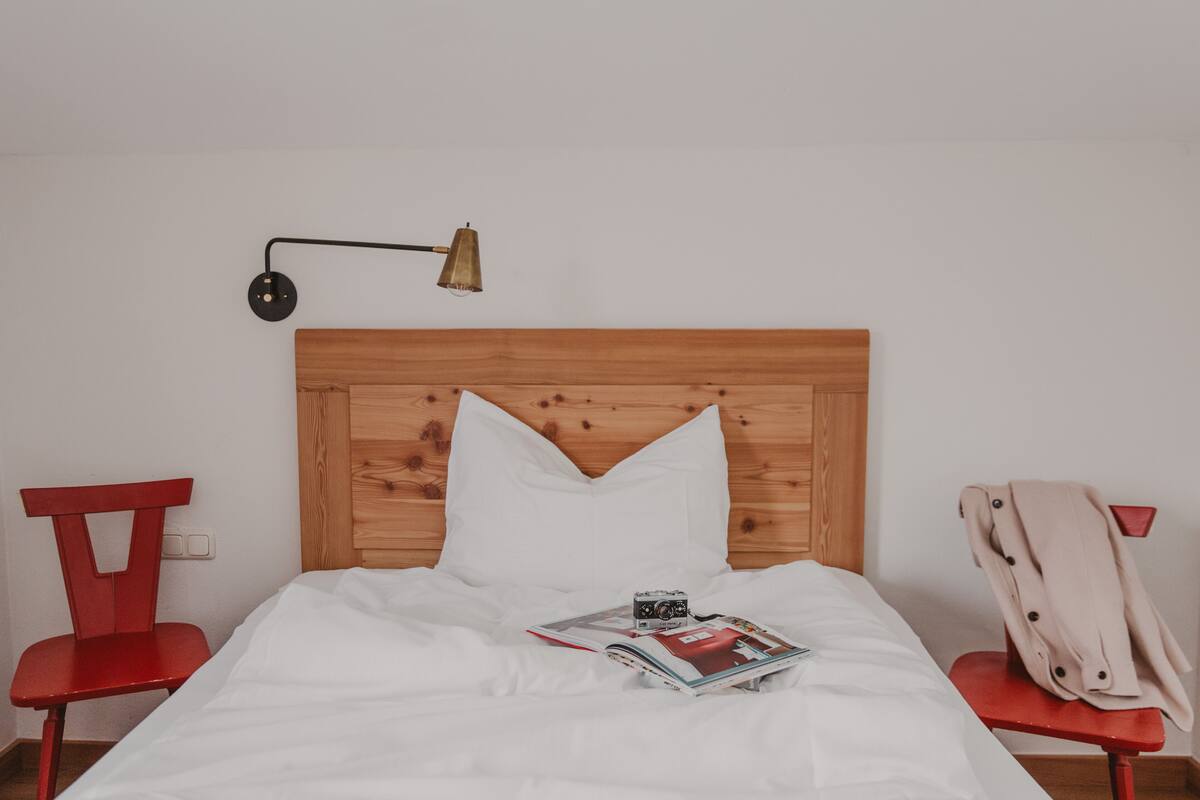 Una cama bien hecha con ropa blanca, una revista y una cámara encima. Hay una silla roja a la izquierda y otra silla roja a la derecha de la cama.