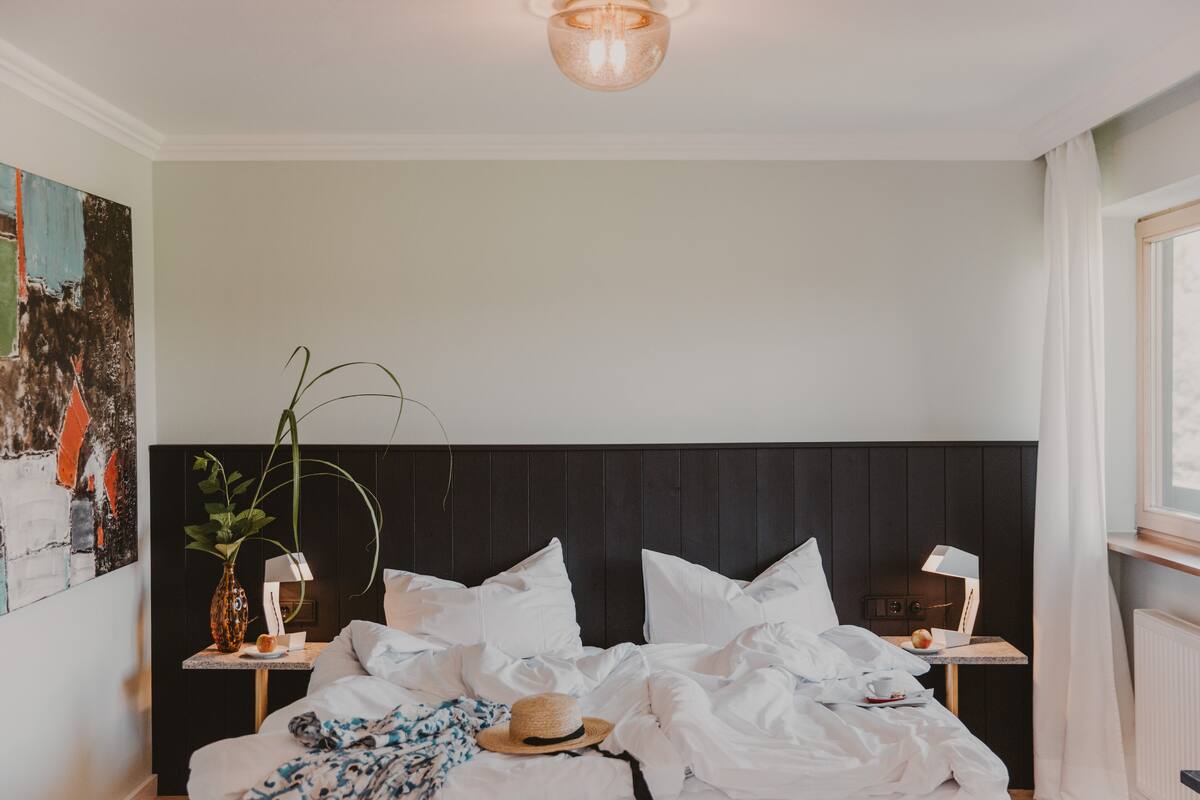 Una cama bien hecha con sábanas blancas, una mesita de noche con lámpara y una planta grande en maceta.