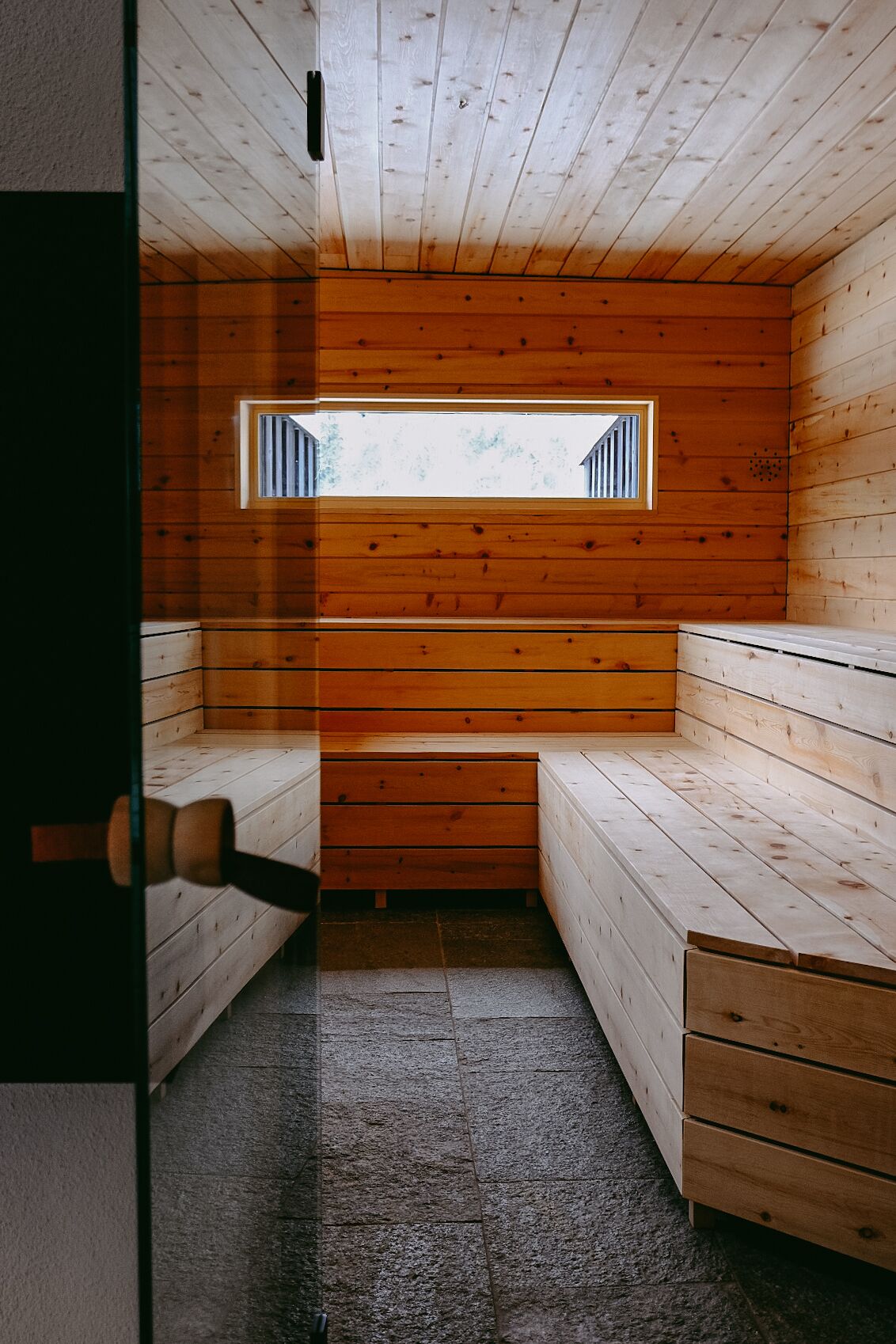 A sauna room with wooden benches, a bench with a window, and a door.