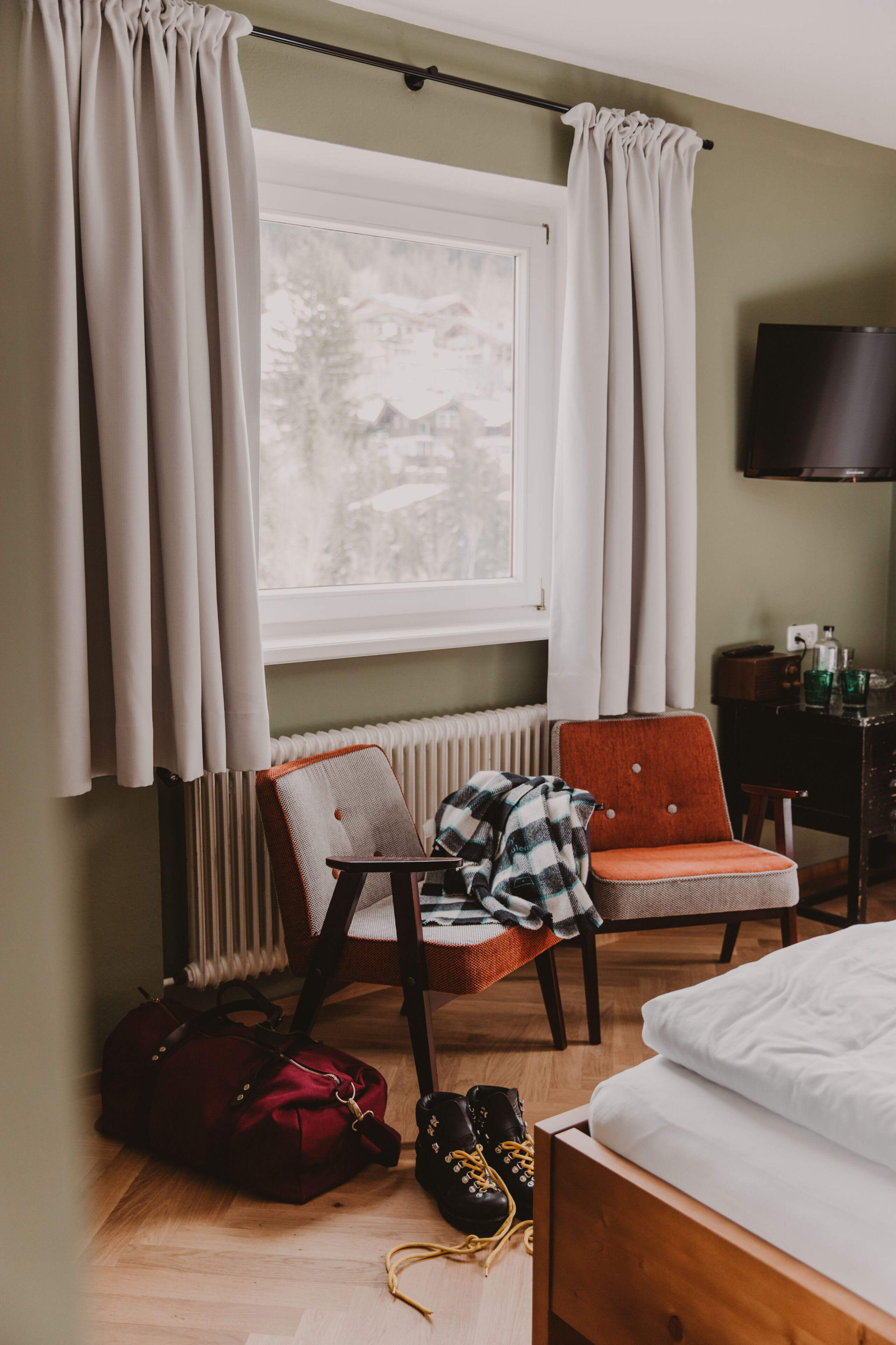 A hotel room with a bed, a chair, a radiator, a television, and a window with curtains.
