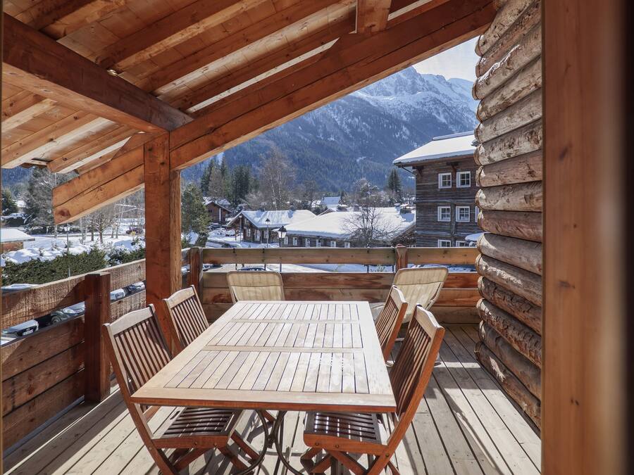 A wooden deck with a table and chairs, overlooking a snowy village and mountains.